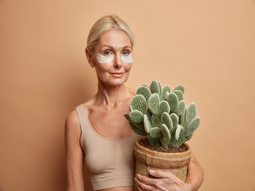 Horizontal shot of good looking middled aged woman has pefect facial skin aplies beauty pads under eyes to remove wrinkles holds pot of cactus isolated over beige background. Body care concept